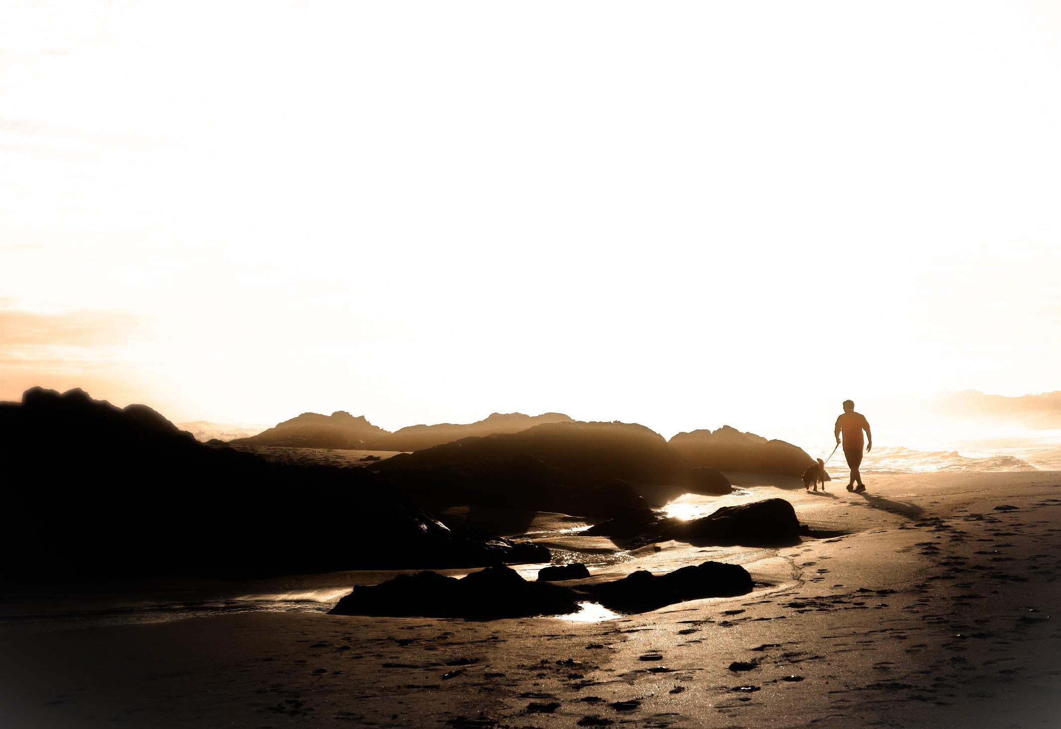 Silhouette of a person walking along a beach at sunset with rocks and water.