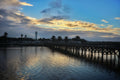 Wooden pier extending into a body of water with a lighthouse in the distance under a cloudy sky.