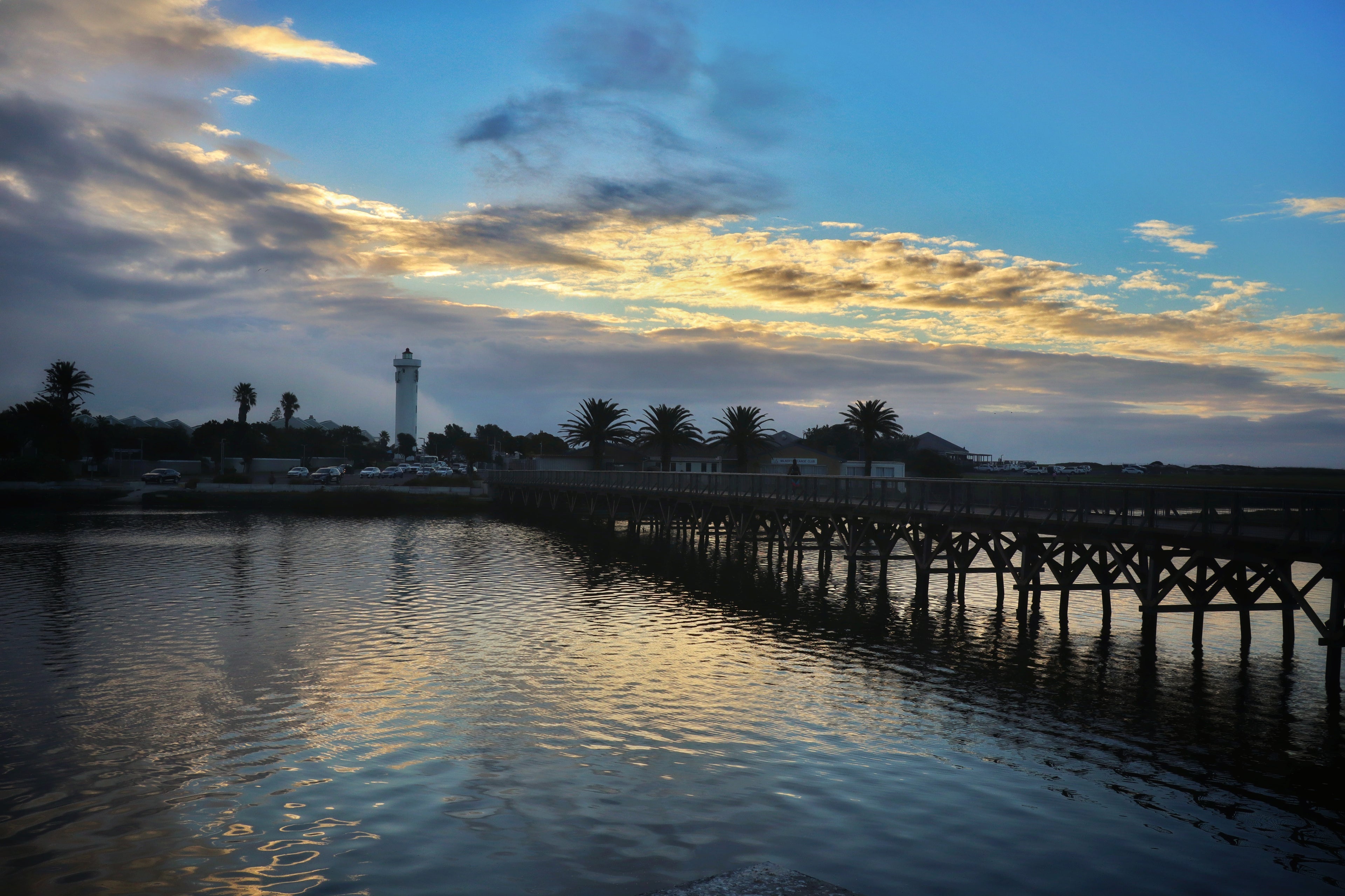 Wooden pier extending into a body of water with a lighthouse in the distance under a cloudy sky.