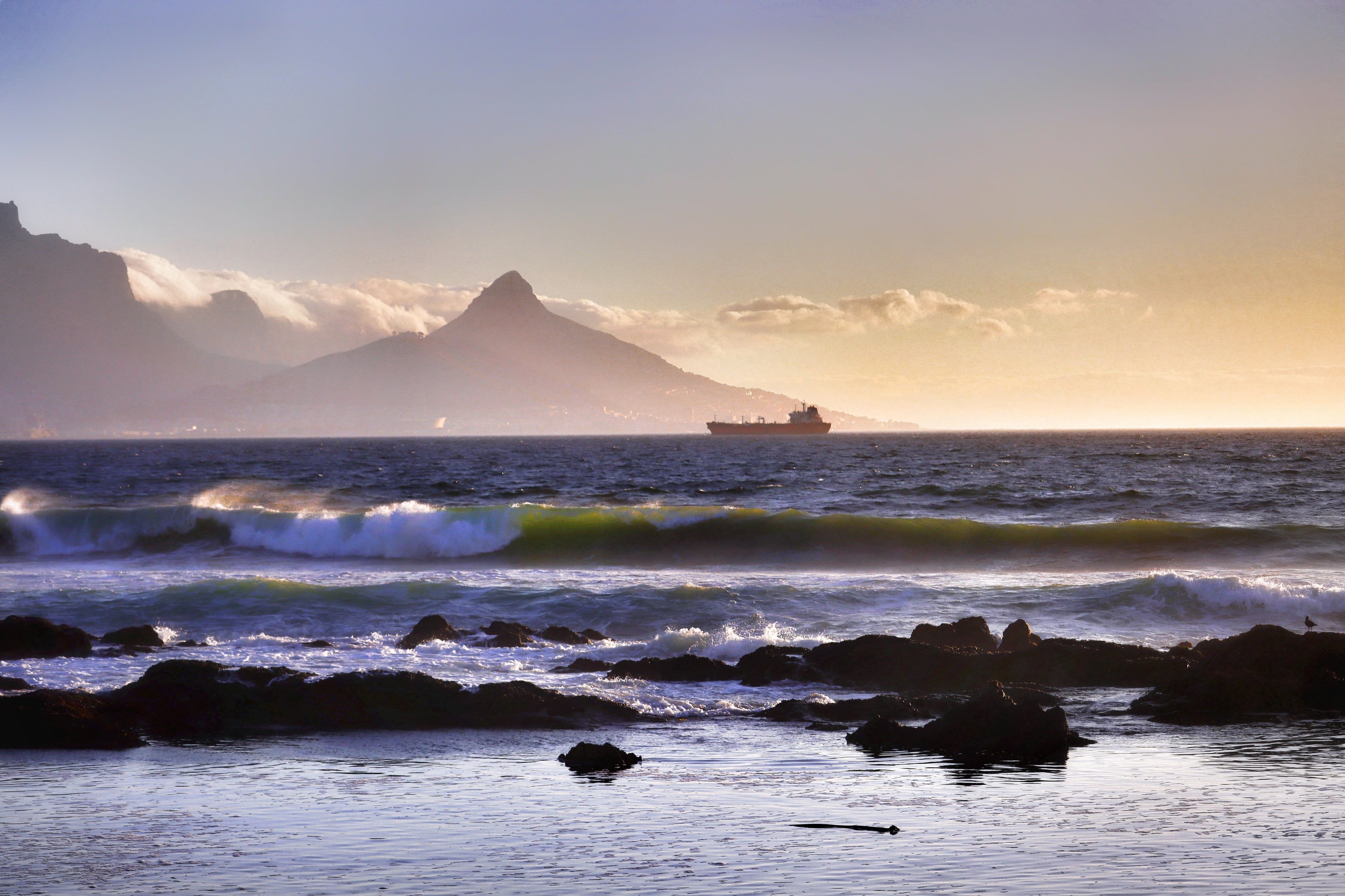 Fine art photograph of golden hour waves breaking over rocks on the Cape Town coast with Table Mountain and Lion's Head silhouetted in warm haze, South African seascape wall art print.