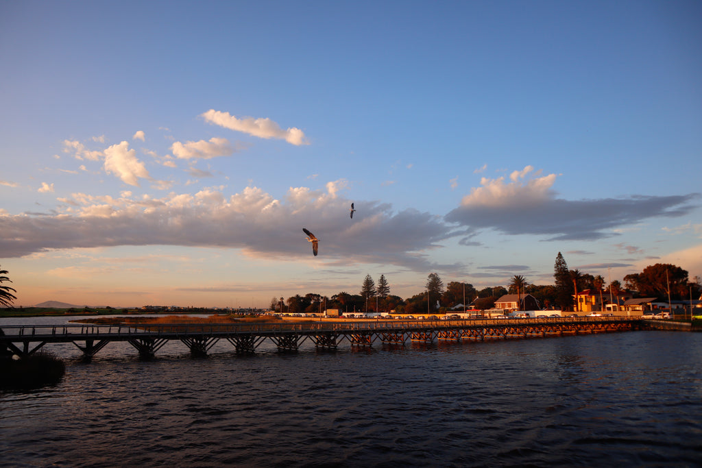 A wide landscape photograph of the historic wooden bridge crossing the lagoon in Milnerton, Cape Town, under a blue twilight sky with flying birds.